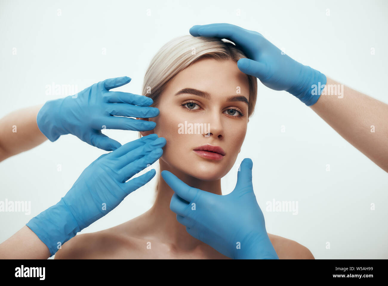 Facial surgery. Beautiful blonde woman waiting for facial surgery while surgeons in blue medical gloves examining her face. Plastic surgery concept. Healthcare. Beauty concept. Stock Photo