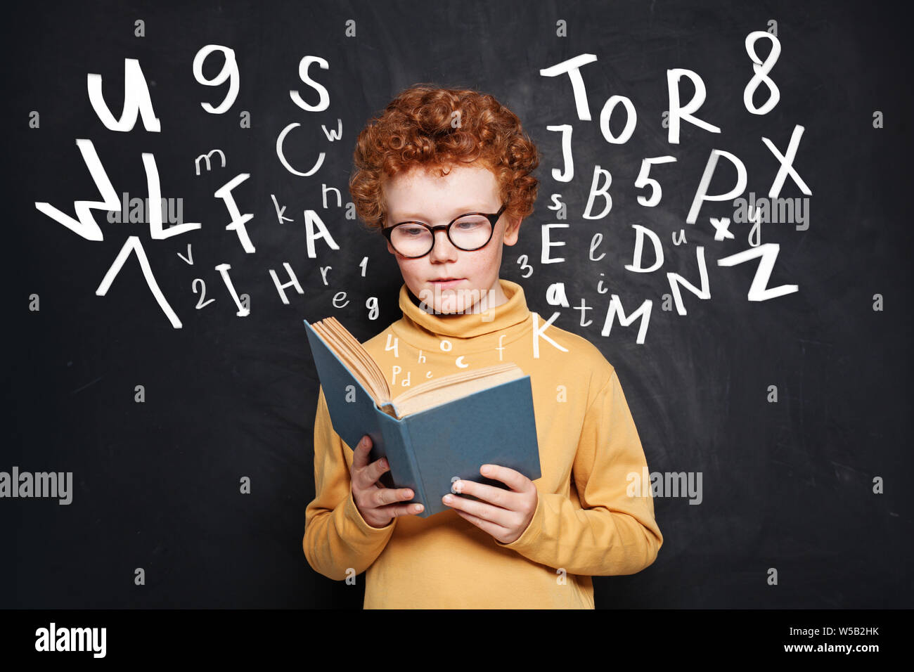 Curious little boy in glasses reading a book against chalkboard Stock Photo