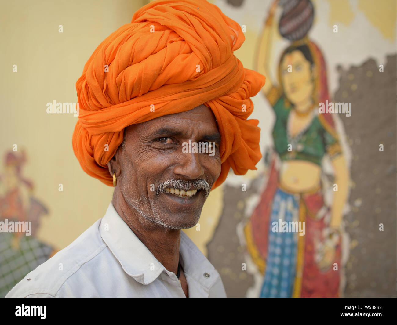 Indian Rajasthani man with an orange Rajasthani turban (pagari) poses for the camera in front of a mural. Stock Photo