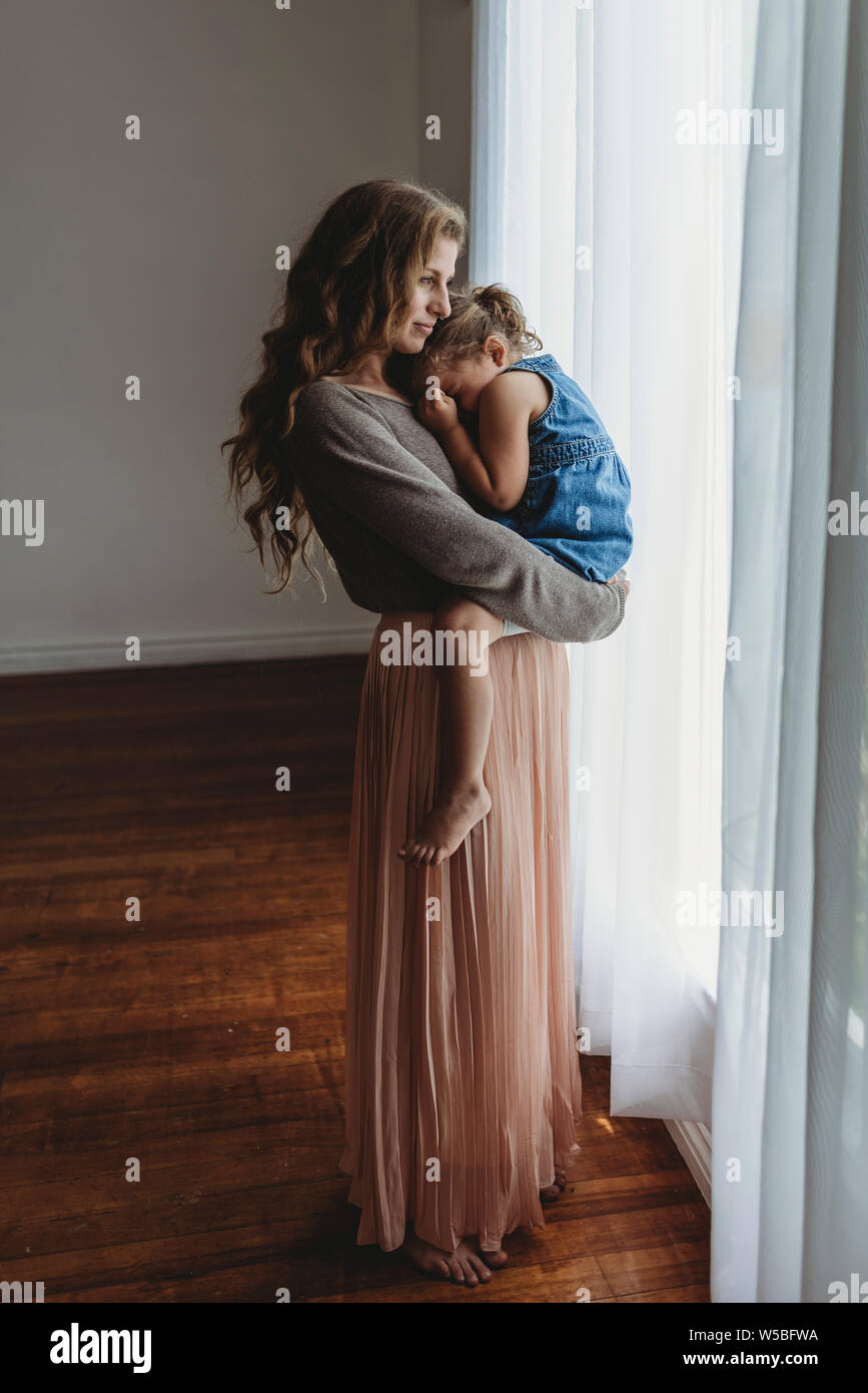 Full length view of mother and daughter cuddling by window in studio ...