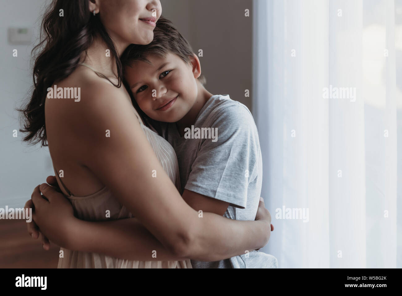 School-aged son being embraced by mother in natural light studio Stock Photo