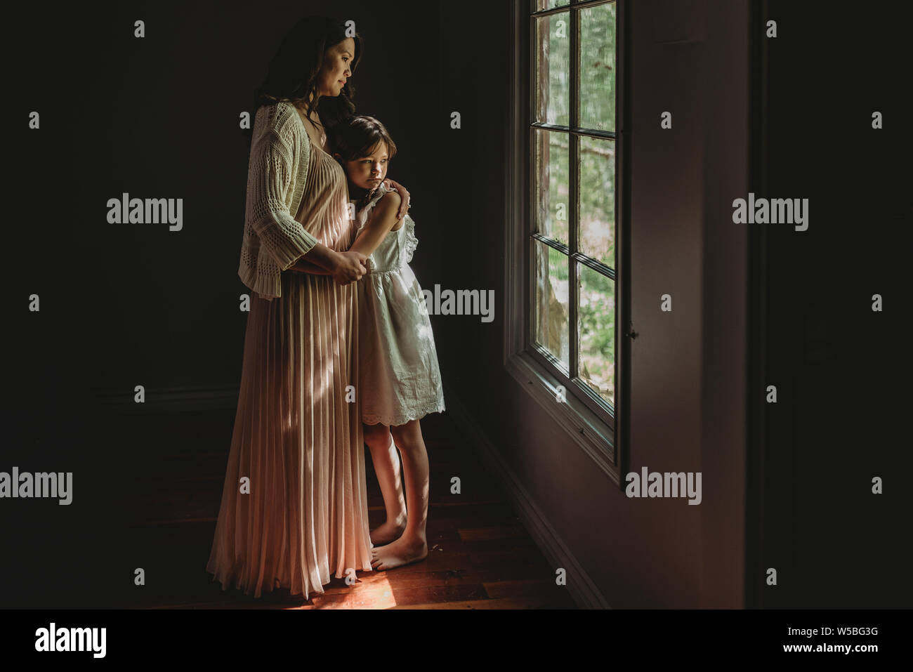 Mother and daughter embrace and looking out studio window Stock Photo