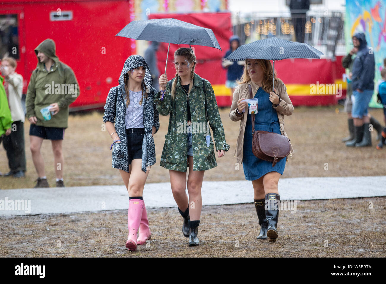 Standon, UK. Saturday 27 July 2019.  A family walking between stages take shelter from the rain under umbrellas at Standon Calling set in the picturesque grounds of Standon Lordship © Jason Richardson / Alamy Live News Stock Photo