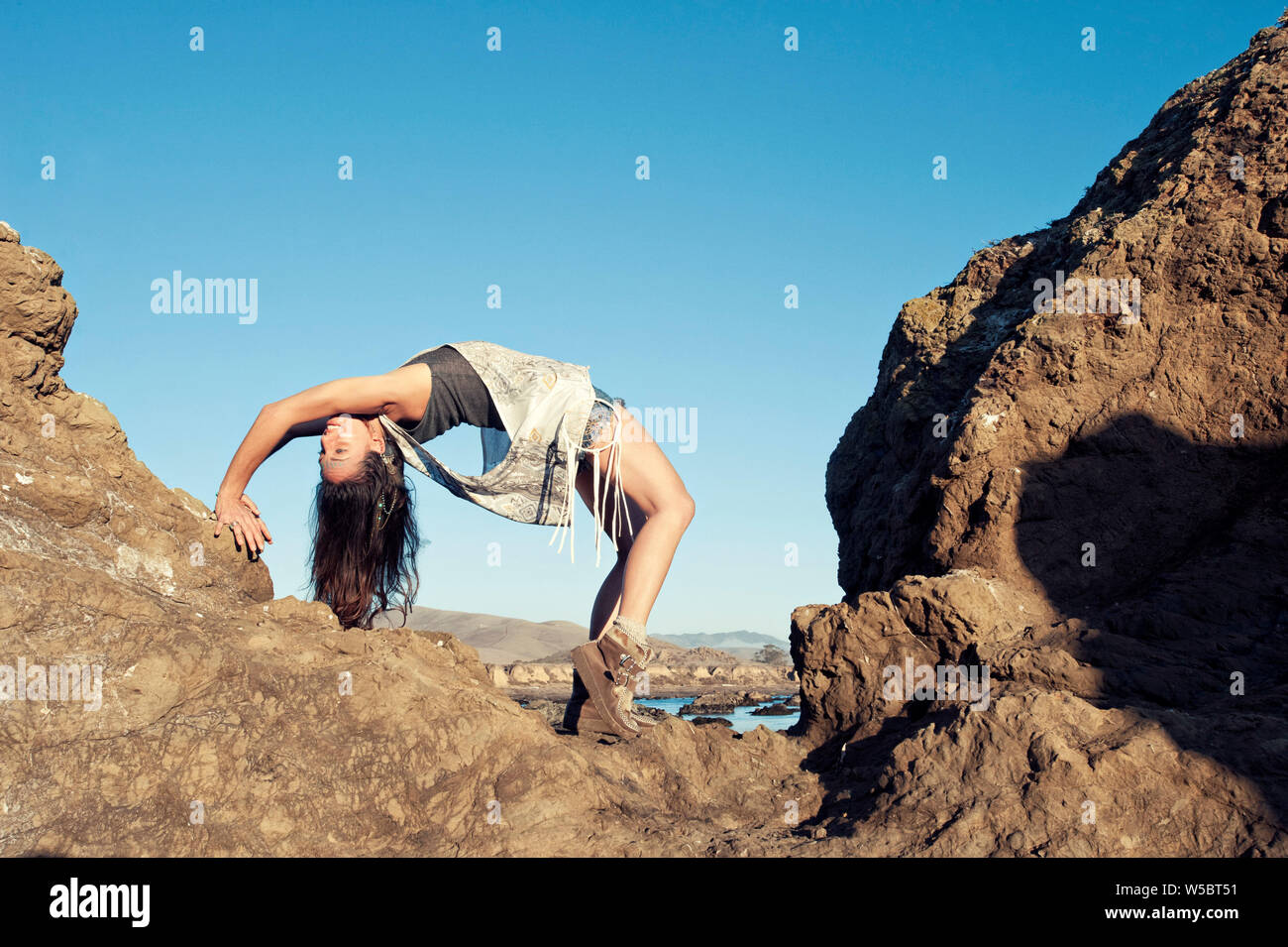 Happy Boho young woman in wheel yoga pose on a rocky wild beach. Stock Photo