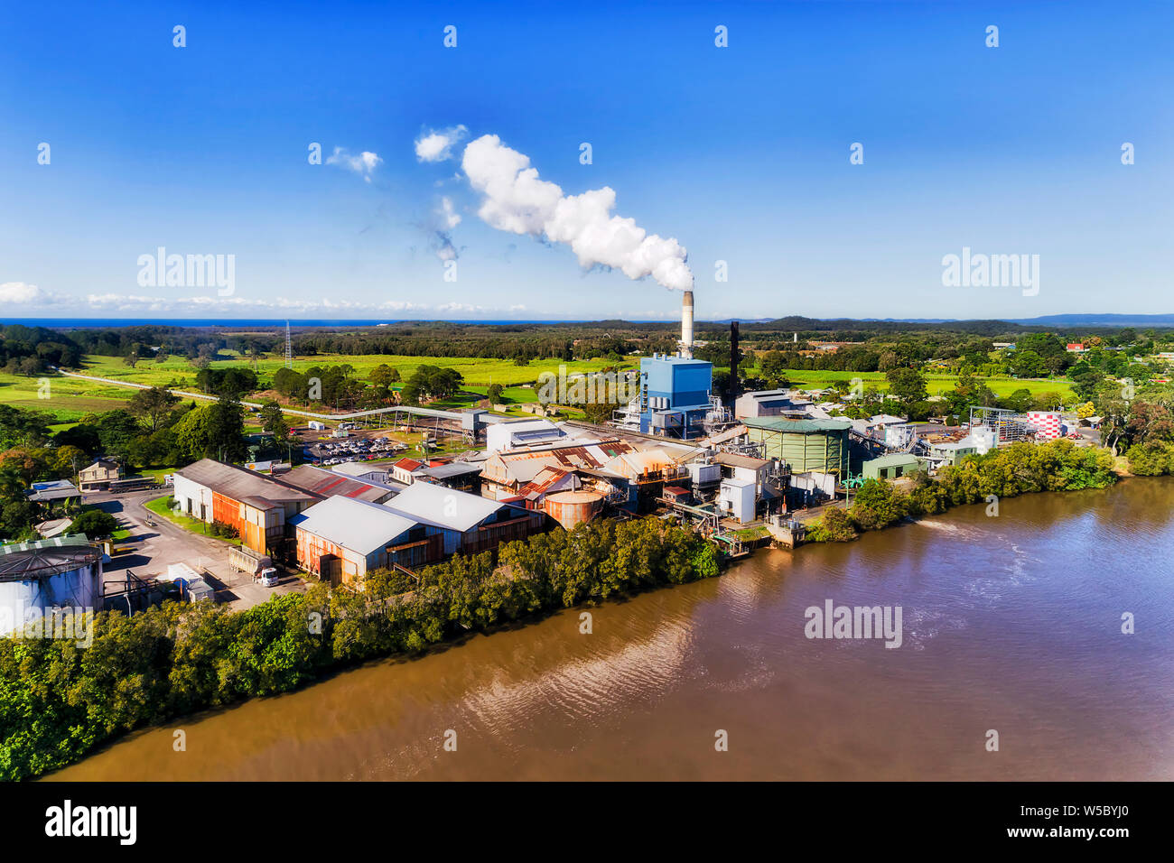 Waterfront of Broadwater sugar mill in rural tropical regional Australia at the centre of sugar cane farms and fields under tall chimney exhausting st Stock Photo