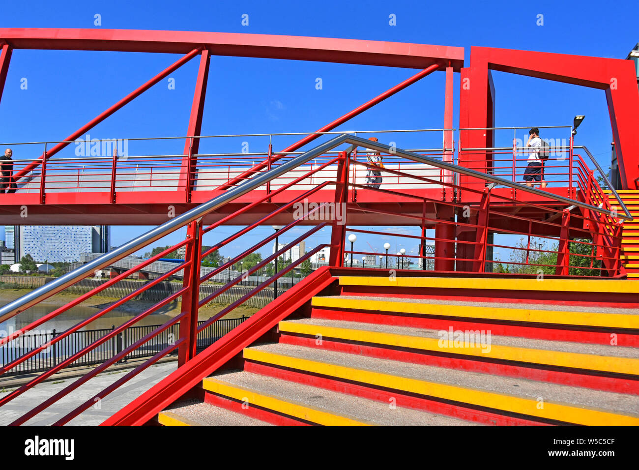Steps & pedestrians at colourful steel footbridge over River Lea links train station to new housing & office development Canning Town East London UK Stock Photo