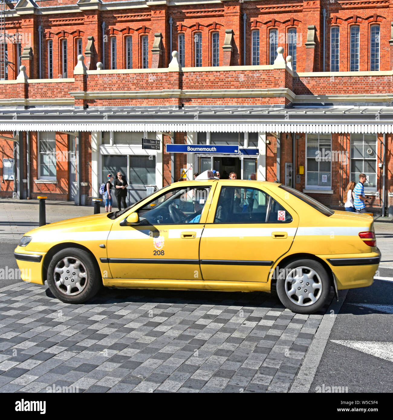 Yellow taxi cab car driver & passenger driving across cobbled speed hump Bournemouth railway train station Victorian brick building Dorset England UK Stock Photo