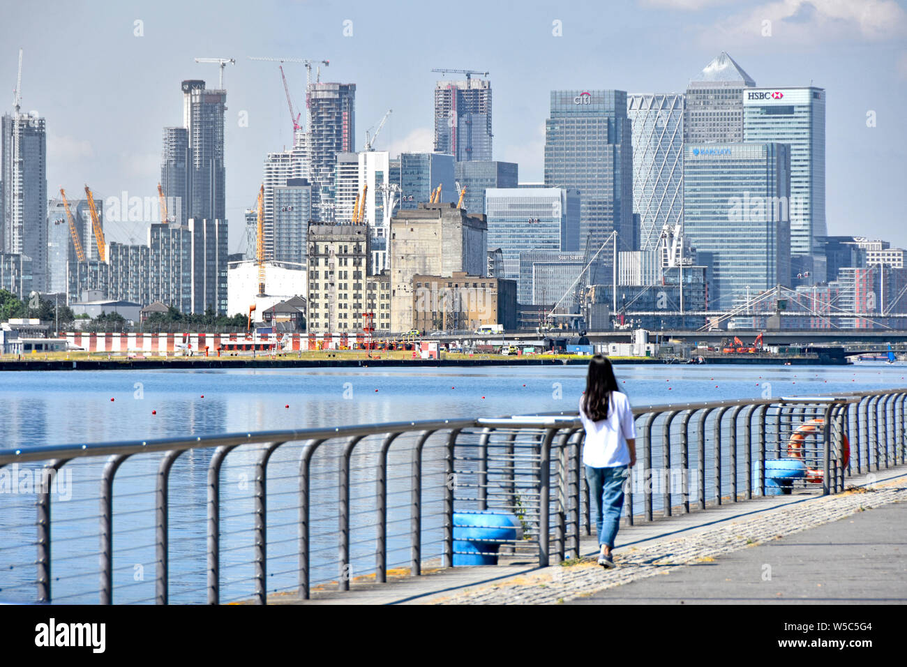 Teenage girl stands beside Royal Docks view looking across water to business development of Canary Wharf cityscape skyline in east London Docklands UK Stock Photo