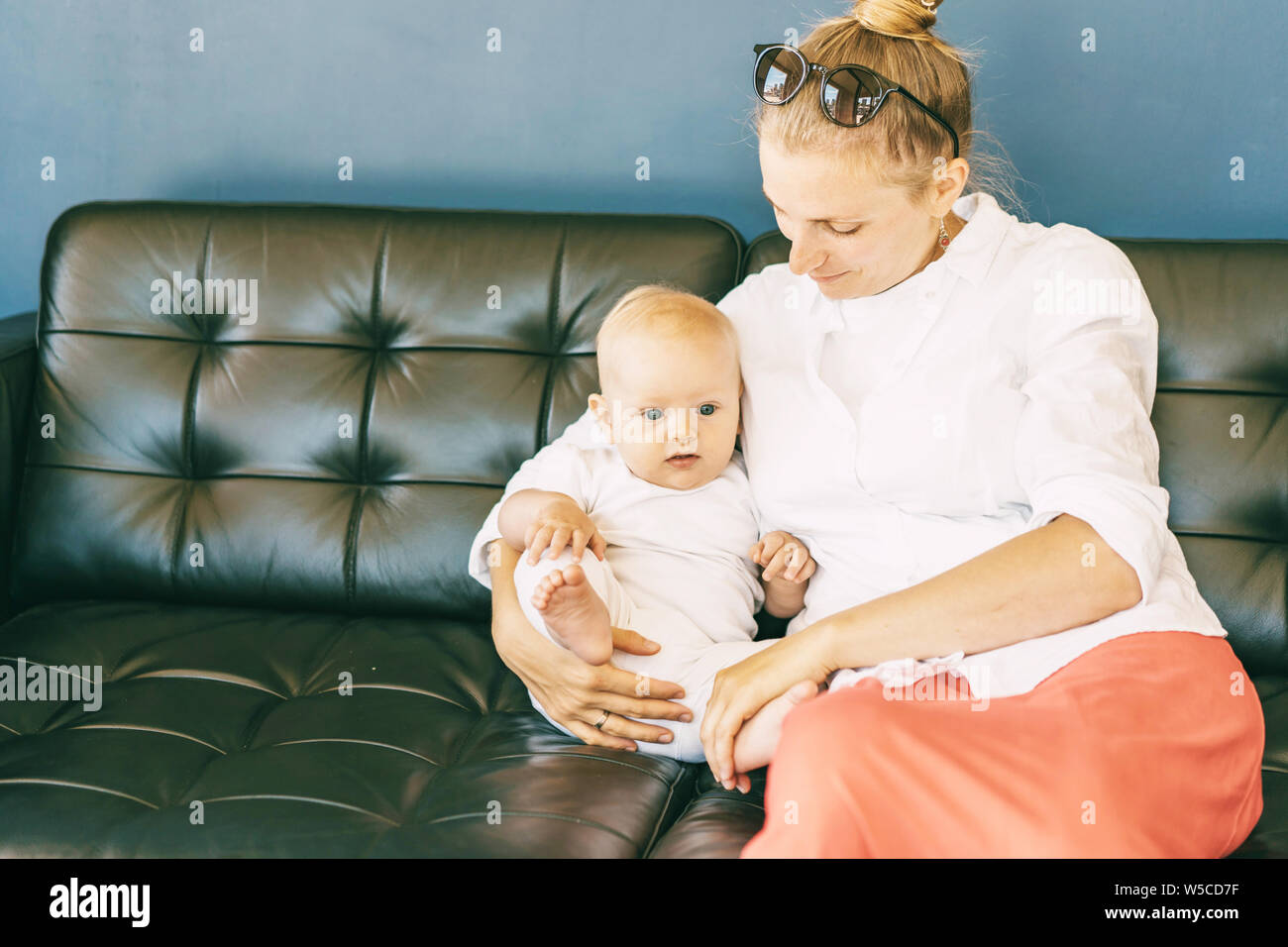 A young mother and a 6-month-old baby are sitting on a dark sofa. Stock Photo
