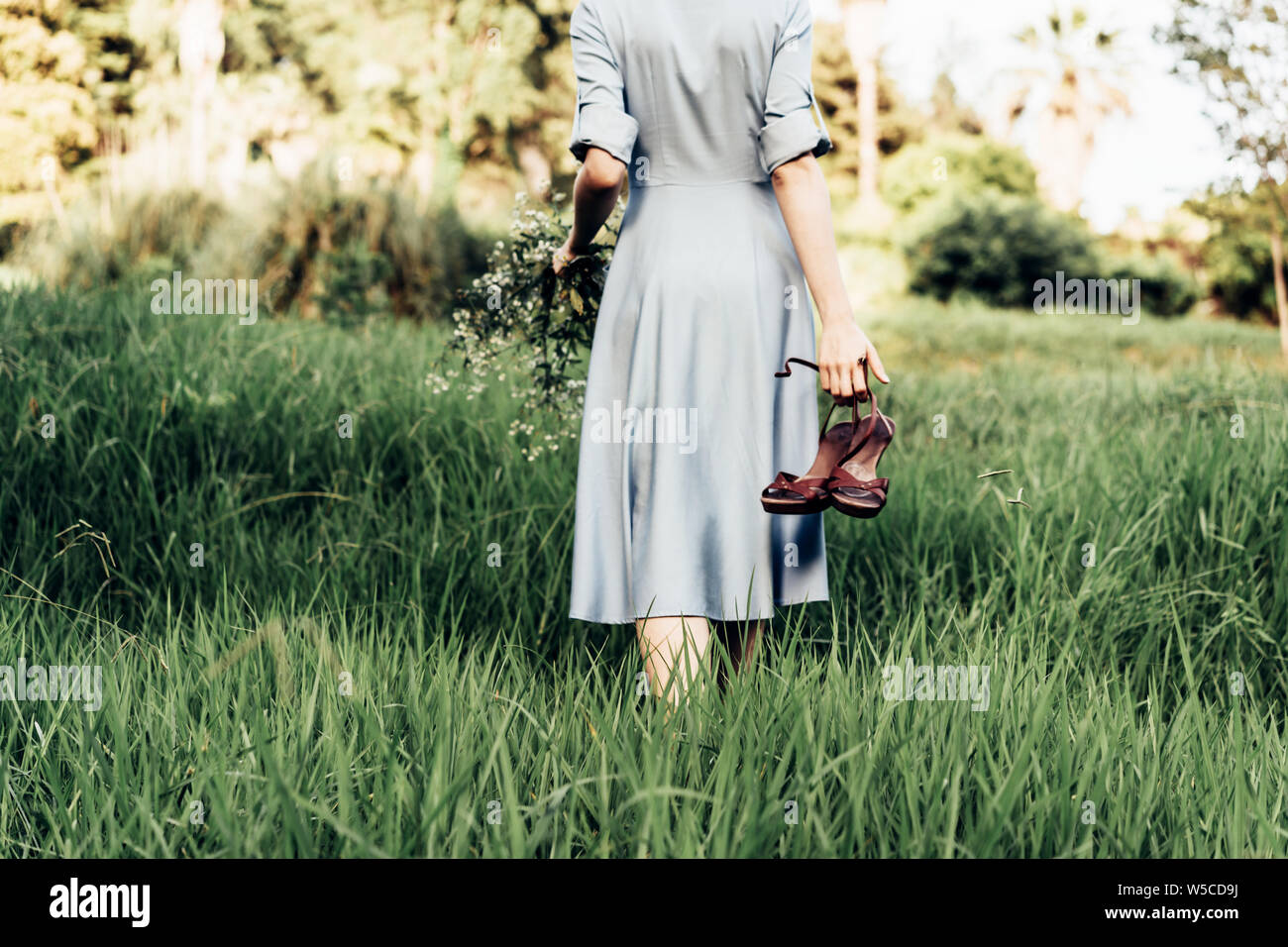 Rear view of a woman in a dress walking along a meadow barefoot with shoes in her hand Stock Photo