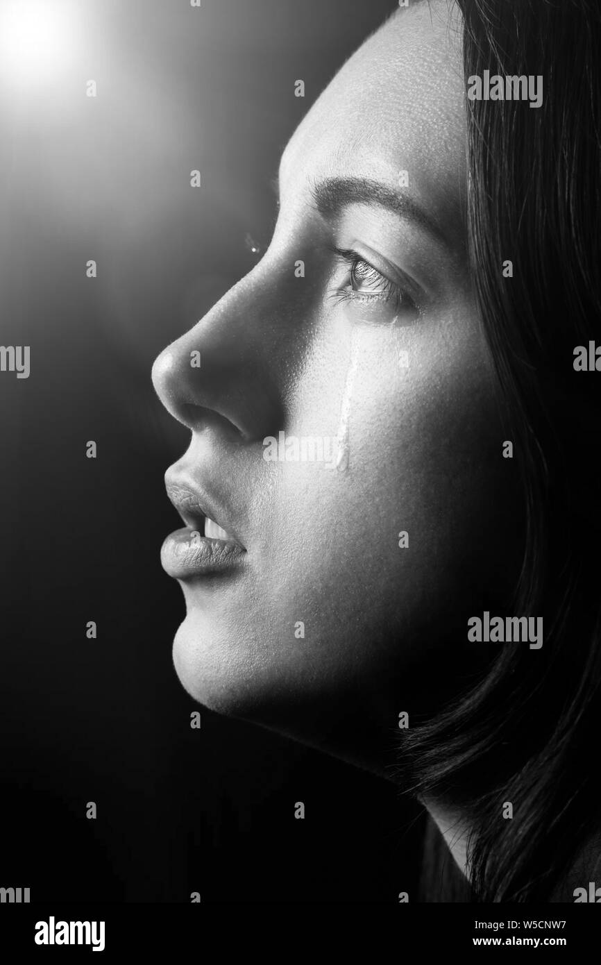 sad woman crying, looking aside on black background with light rays, closeup portrait, profile view, monochrome Stock Photo