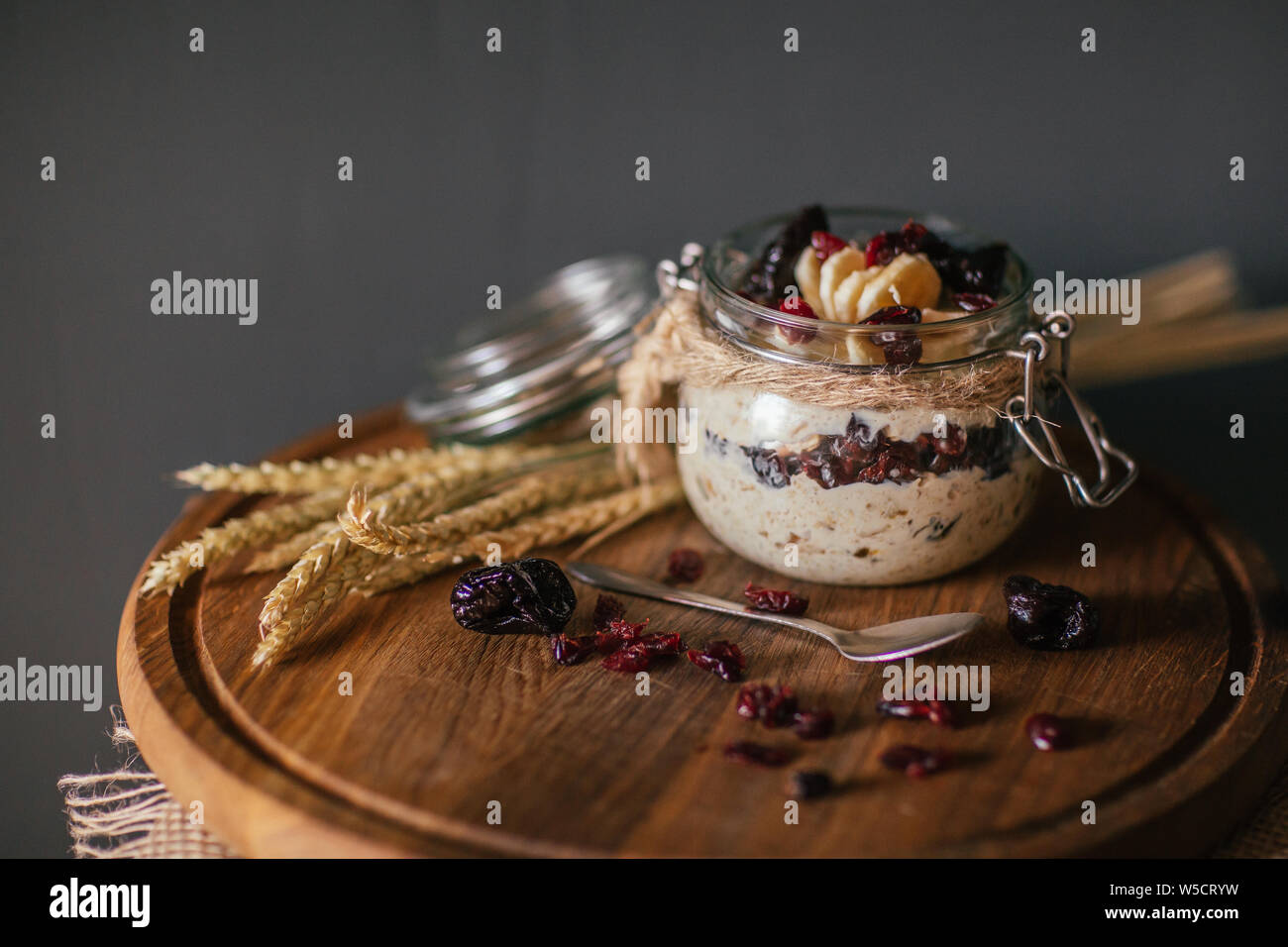 Lazy overnight oatmeal in glass transparent jars with cherry and bananas on a dark black background on a wooden plate decorated with spikelets of whea Stock Photo