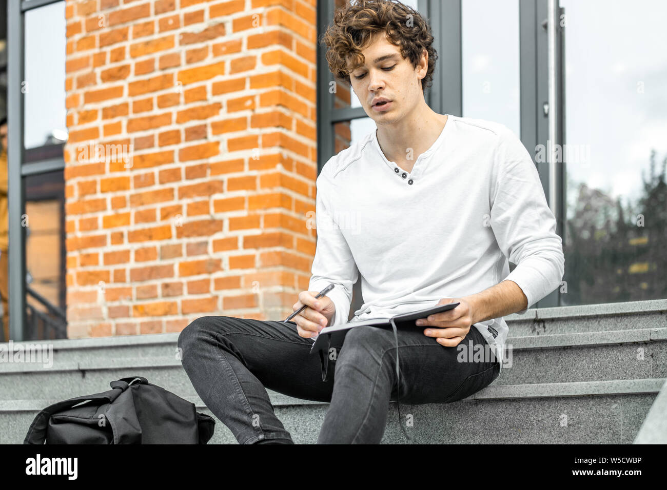 Attractive man holding notepad in his hands and sitting on concrete steps of red bricked building. Stock Photo