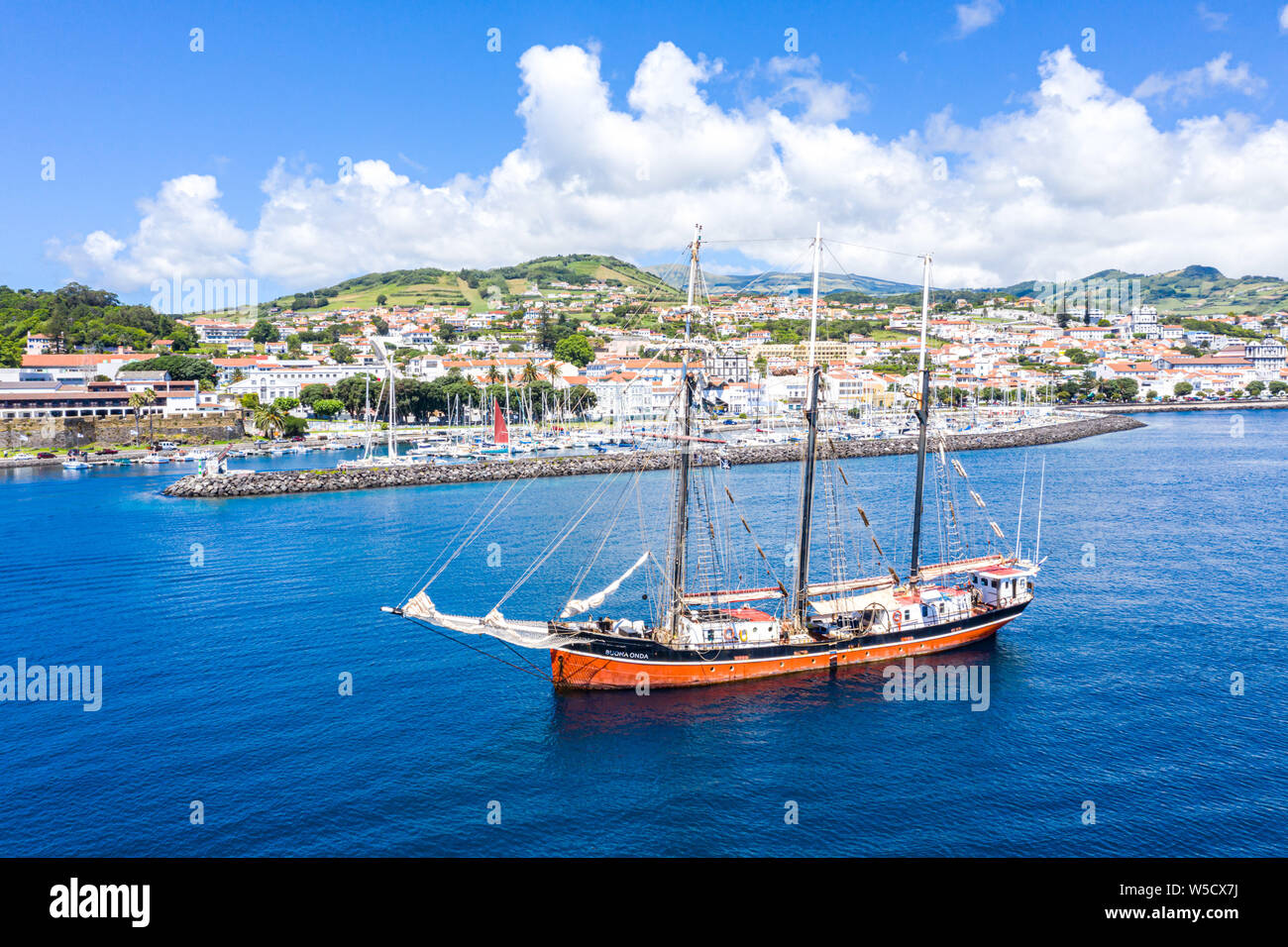 Horta, Portugal - Jul 16 2019: Aerial view of three masted schooner Sailing Vessel Buona Onda on roadstead, historical touristic hilly town centre and Stock Photo