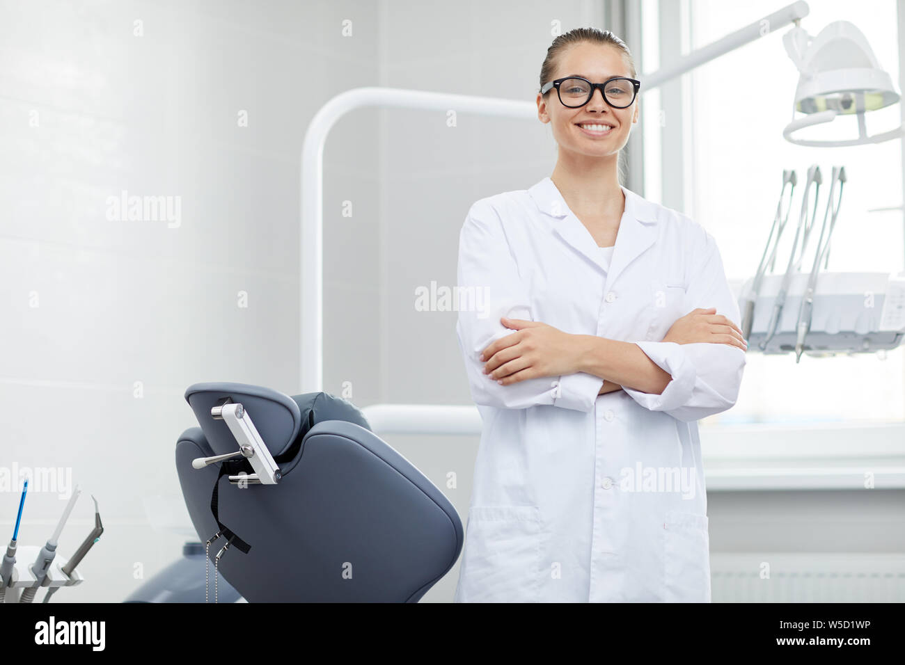 Portrait of young female doctor wearing glasses smiling at camera while posing with dental chair
