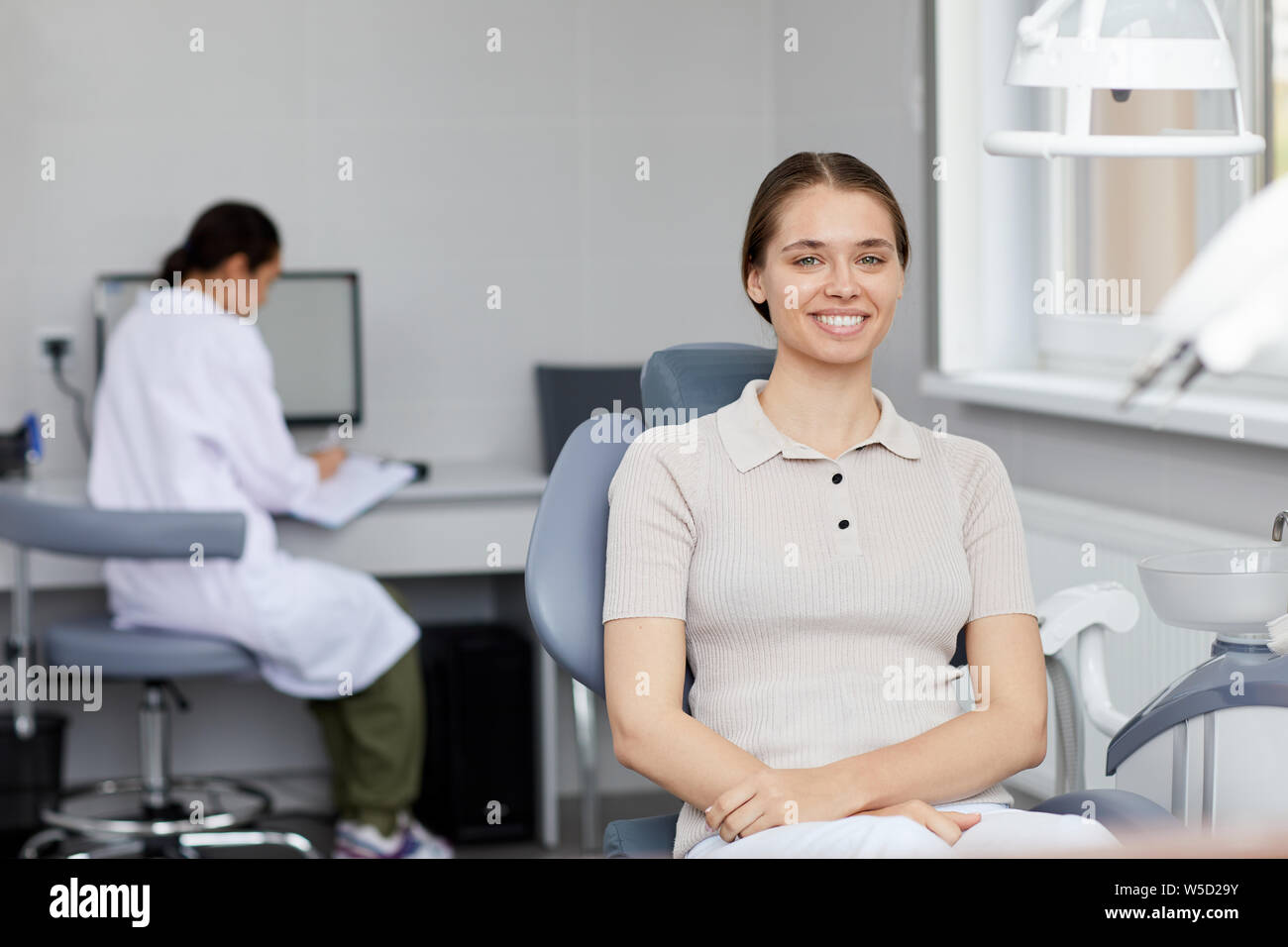 Portrait of pretty young woman smiling at camera and showing perfect white teeth while sitting in dental chair and waiting for appointment, copy space Stock Photo