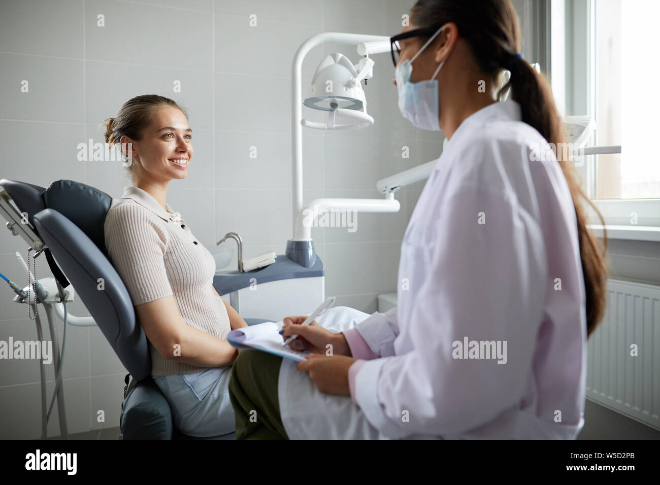 Side view portrait of pretty young woman sitting in dental chair and smiling at doctor during consultation in clinic, copy space Stock Photo