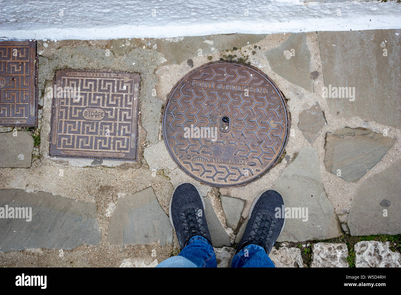 IOANNINA, GREECE - JUNE 6, 2019 - Male feet in blue dusty sneakers positioned next to few manholes at Ioannina Island on lake Pamvotida near the beaut Stock Photo