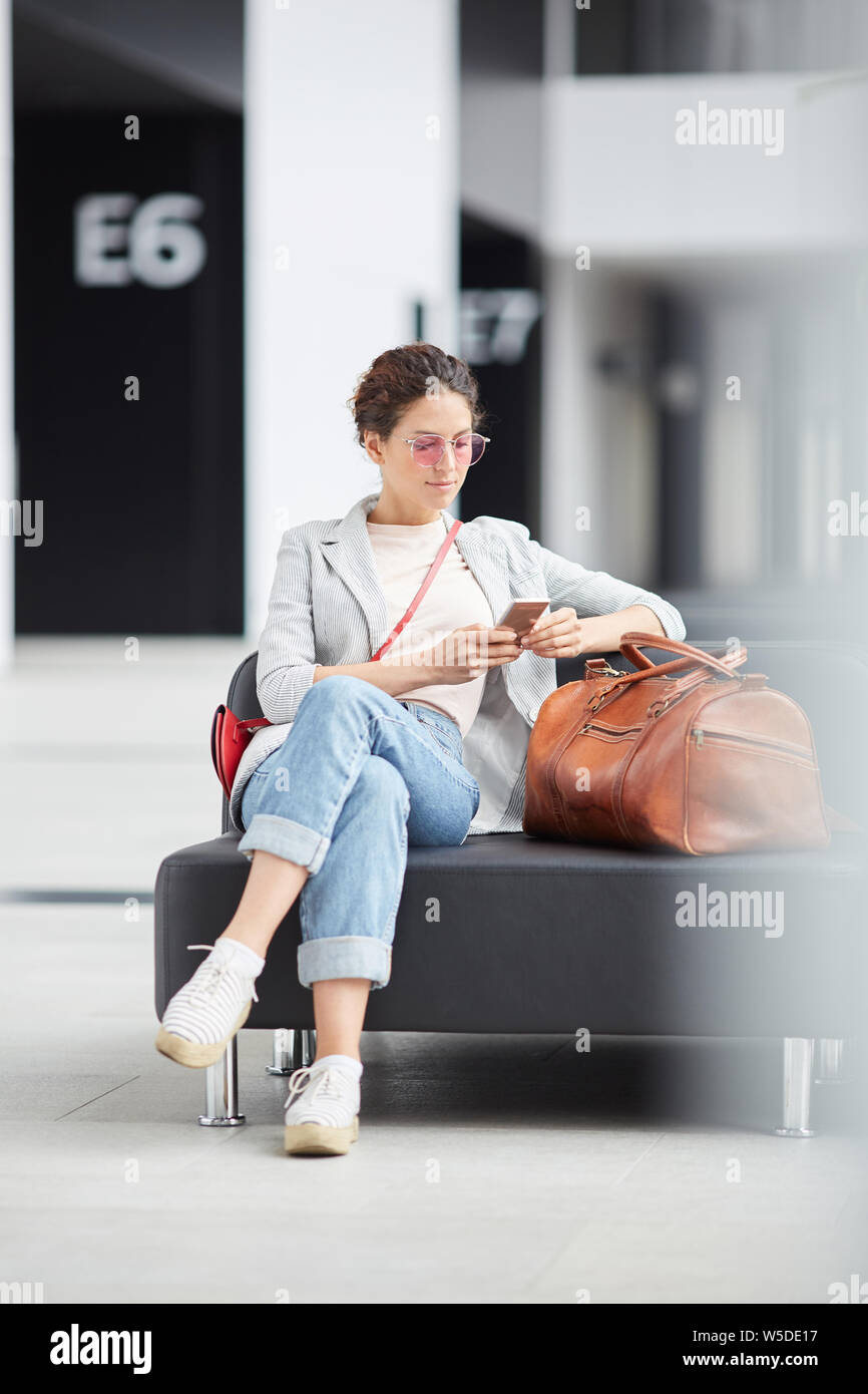 Serious hipster girl in casual outfit sitting on leather sofa with travel bag in airport waiting area and surfing net on phone Stock Photo