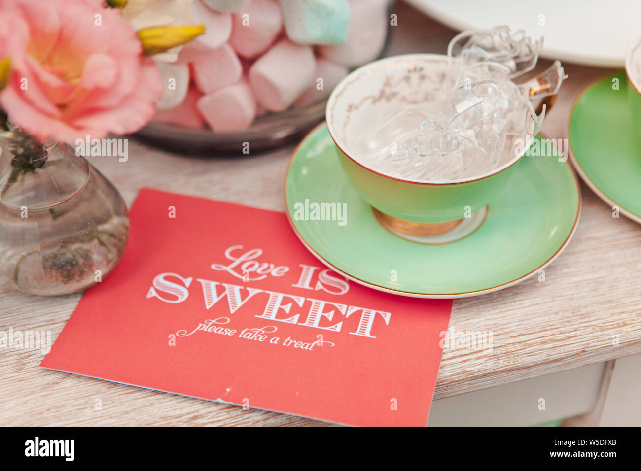 Closeup of a cup with sweets on a background of a vase with marshmallows and a card with the words Love is sweet, selective focus Stock Photo