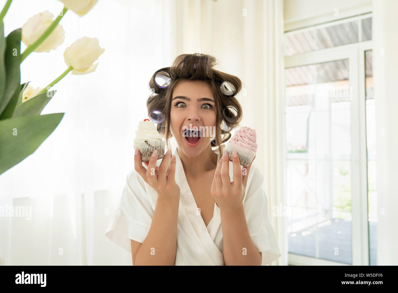 beautiful young brunette woman in hair curlers holding two cupcakes in both hands standing in bright kitchen looking amazed Stock Photo