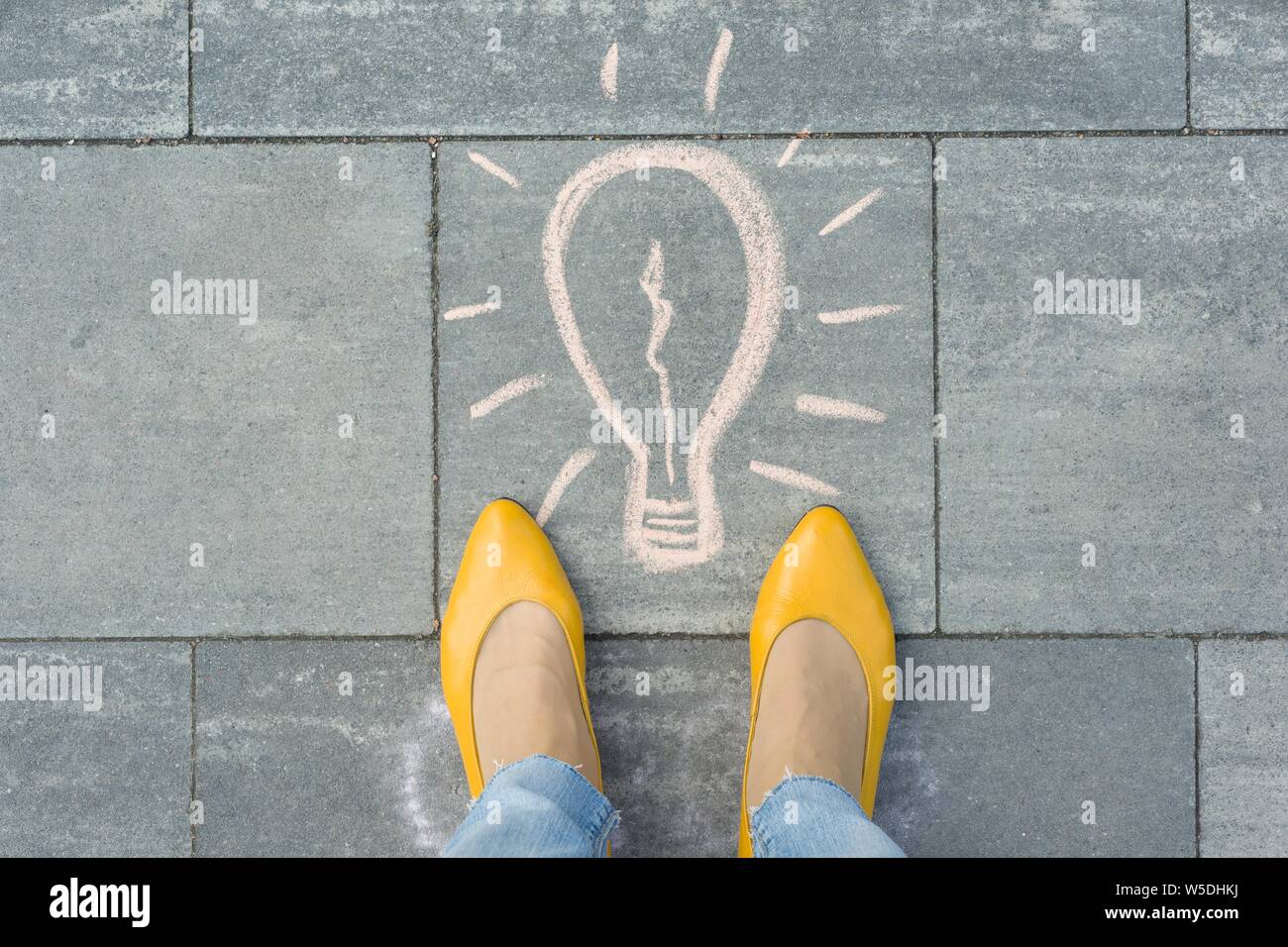 Female feet with abstract image drawing of light bulb written on grey sidewalk. Stock Photo