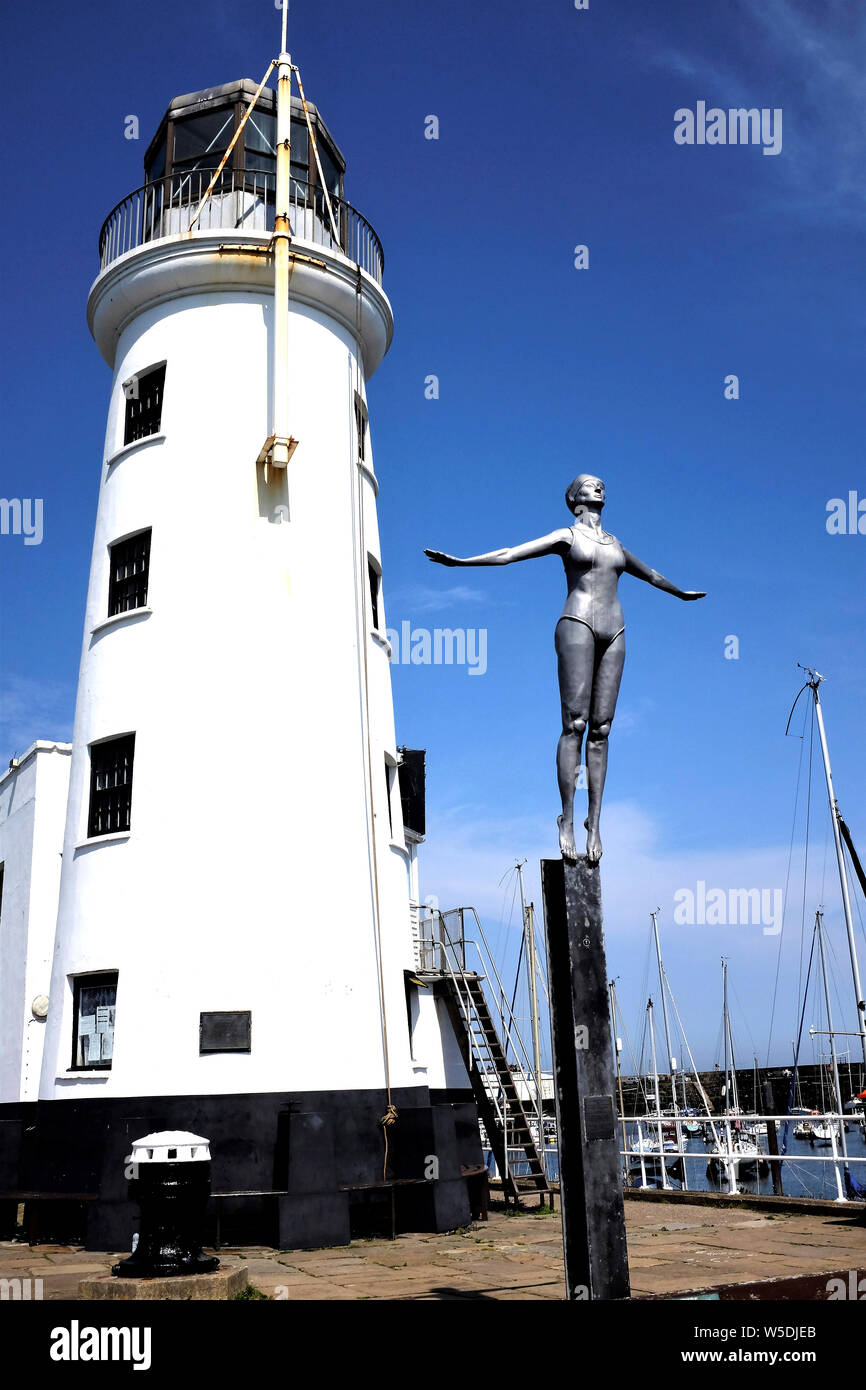 Scarborough, Yorkshire, UK. July 25, 2019. Galvanised steel statue of a diving belle against the lighthouse on the end of Vincent's pier . Stock Photo