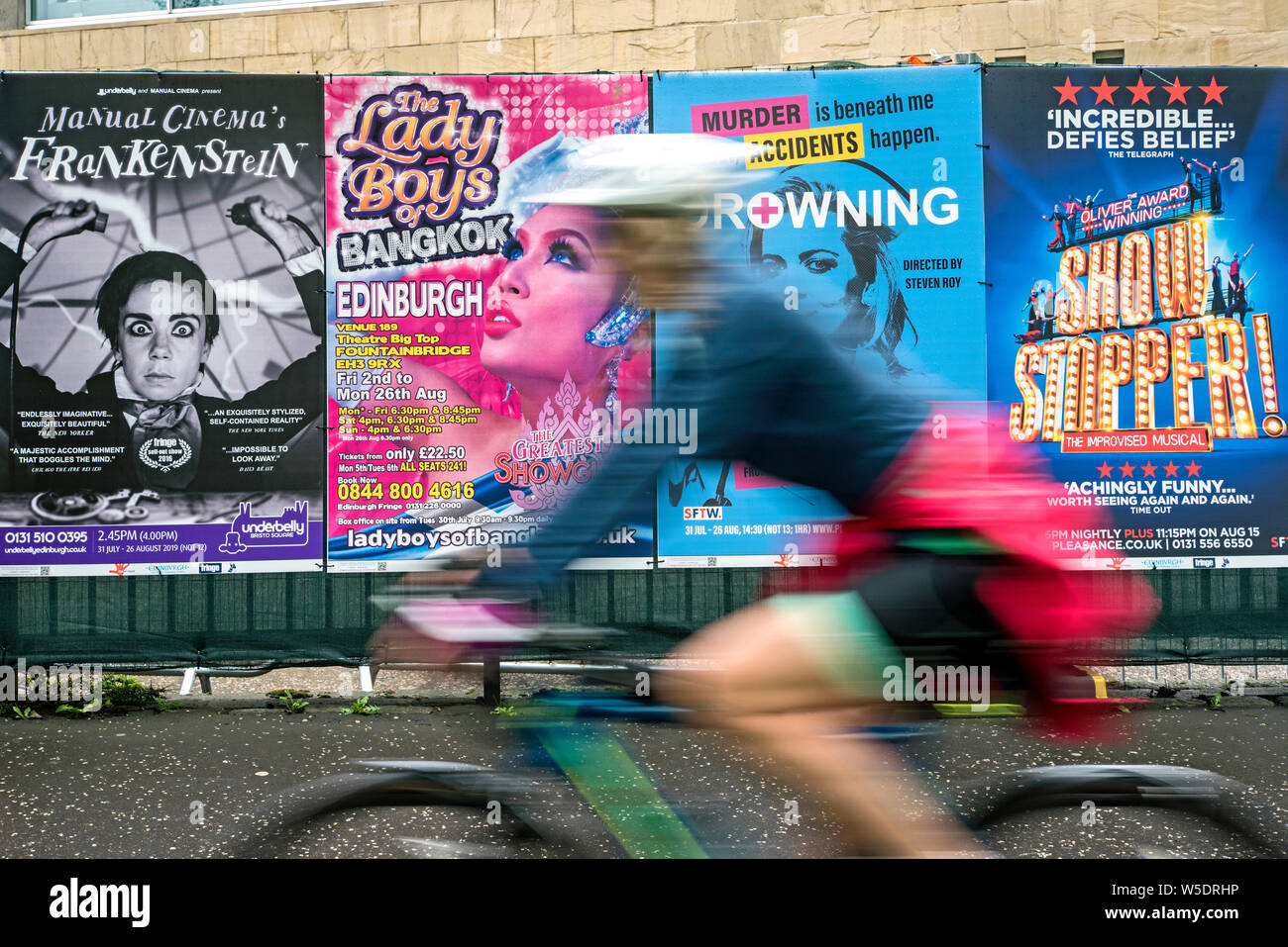A female cyclist rides by advertising posters for Edinburgh Fringe Festival shows in the Meadows, Edinburgh, Scotland, UK. (with motion blur) Stock Photo