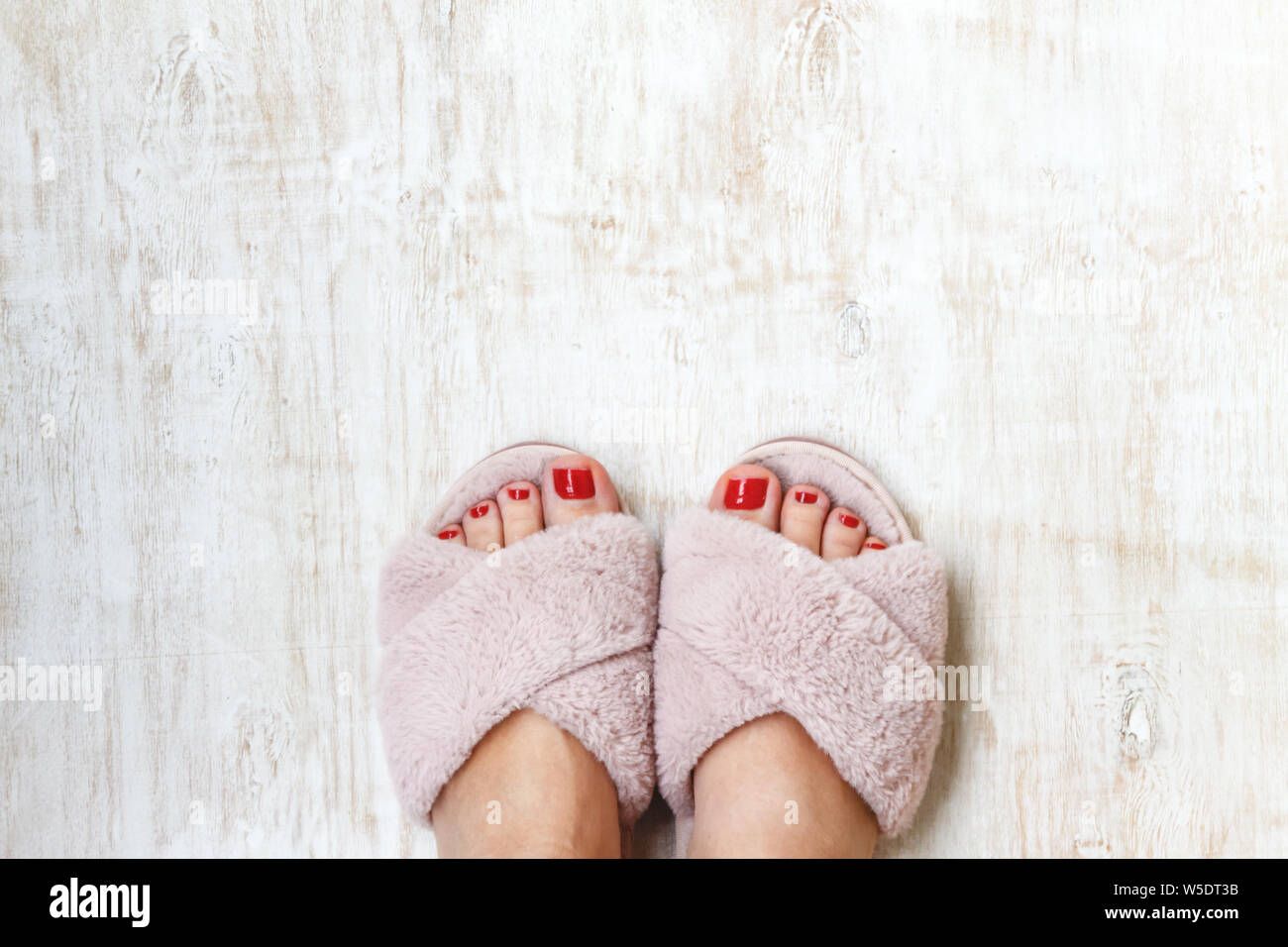 female legs with red nails in home fur fluffy pink slippers on a light wooden background. flat lay. Top view. The concept of a cozy bright girl house. Stock Photo