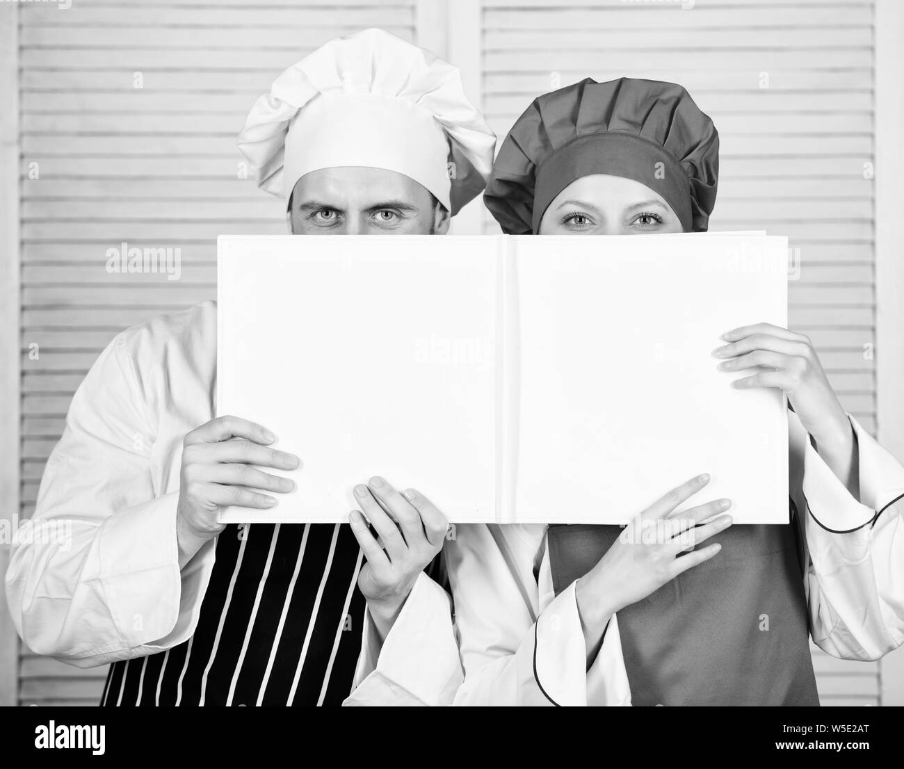 Book family recipes. Cooking guide. According to recipe. Man and woman chef hide faces behind open book. Guy and girl read book recipes. Culinary concept. Family learn recipe. Improve cooking skill. Stock Photo