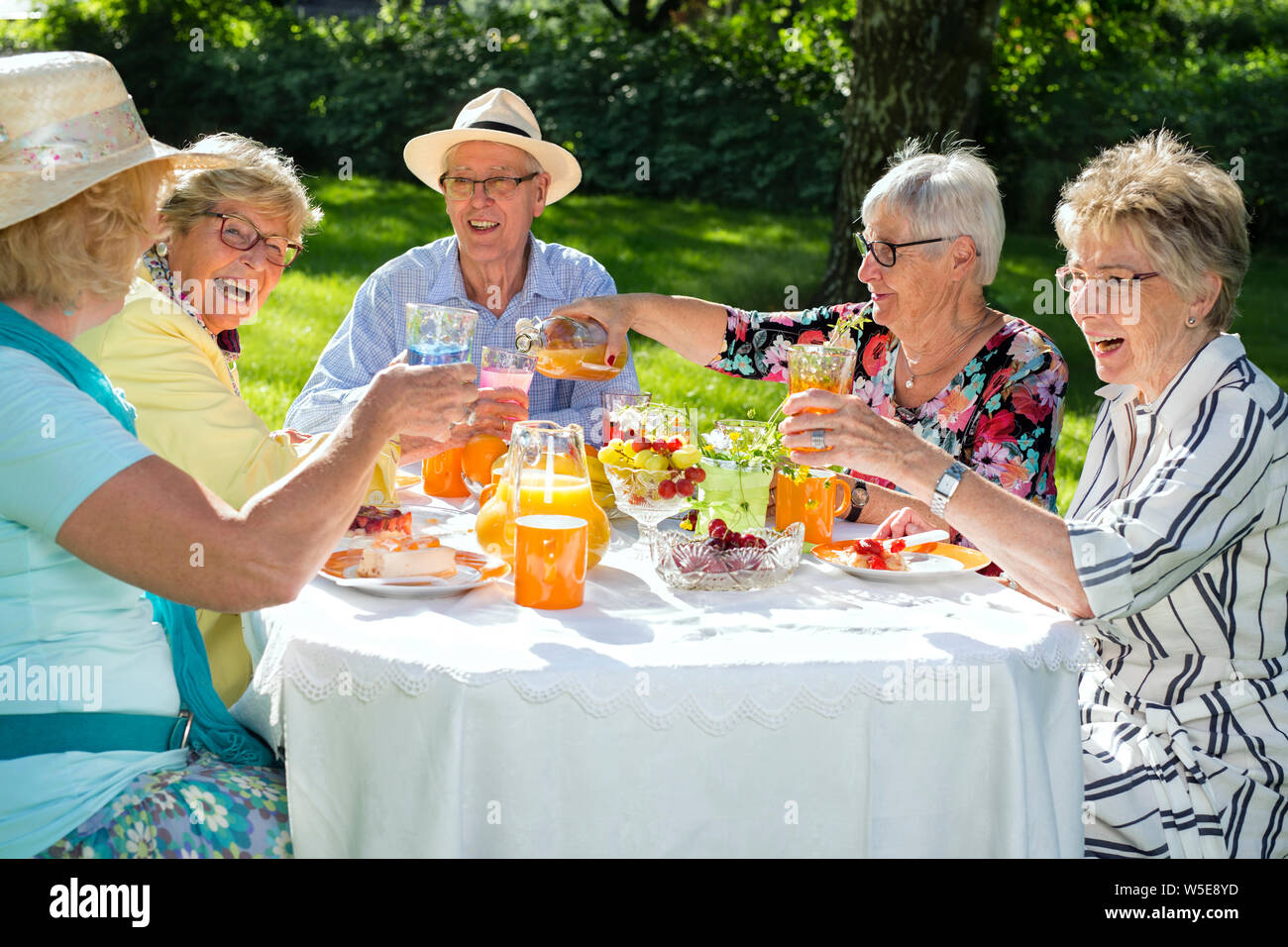 Happy elderly people sitting around the table picnicking. Group of seniors eating fruit cake and drinking orange juice, one woman is serving juice. Stock Photo