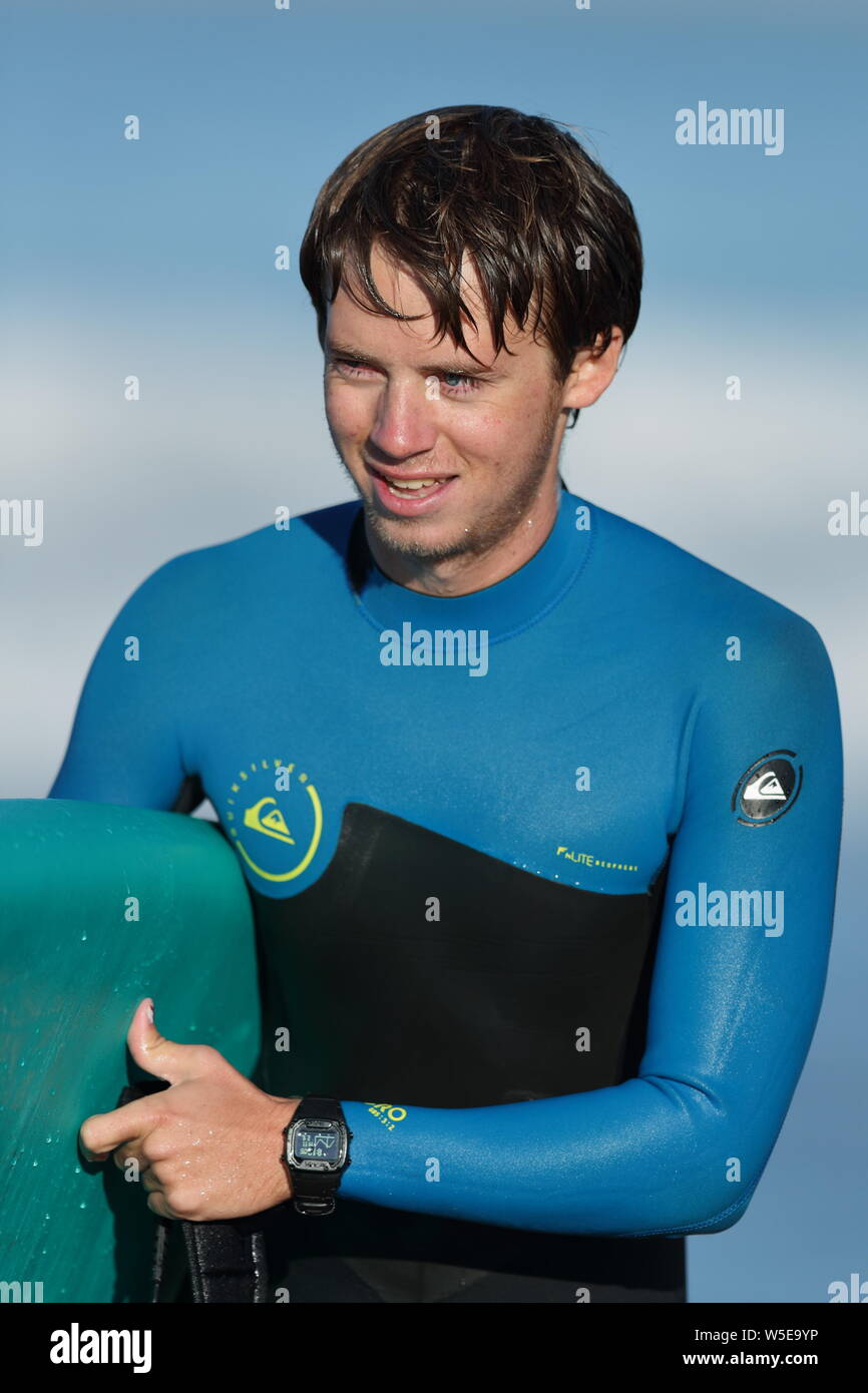 Young man wearing a black and blue wetsuit carrying a blue surfboard in Huntington Beach, California on October 19, 2018 Stock Photo