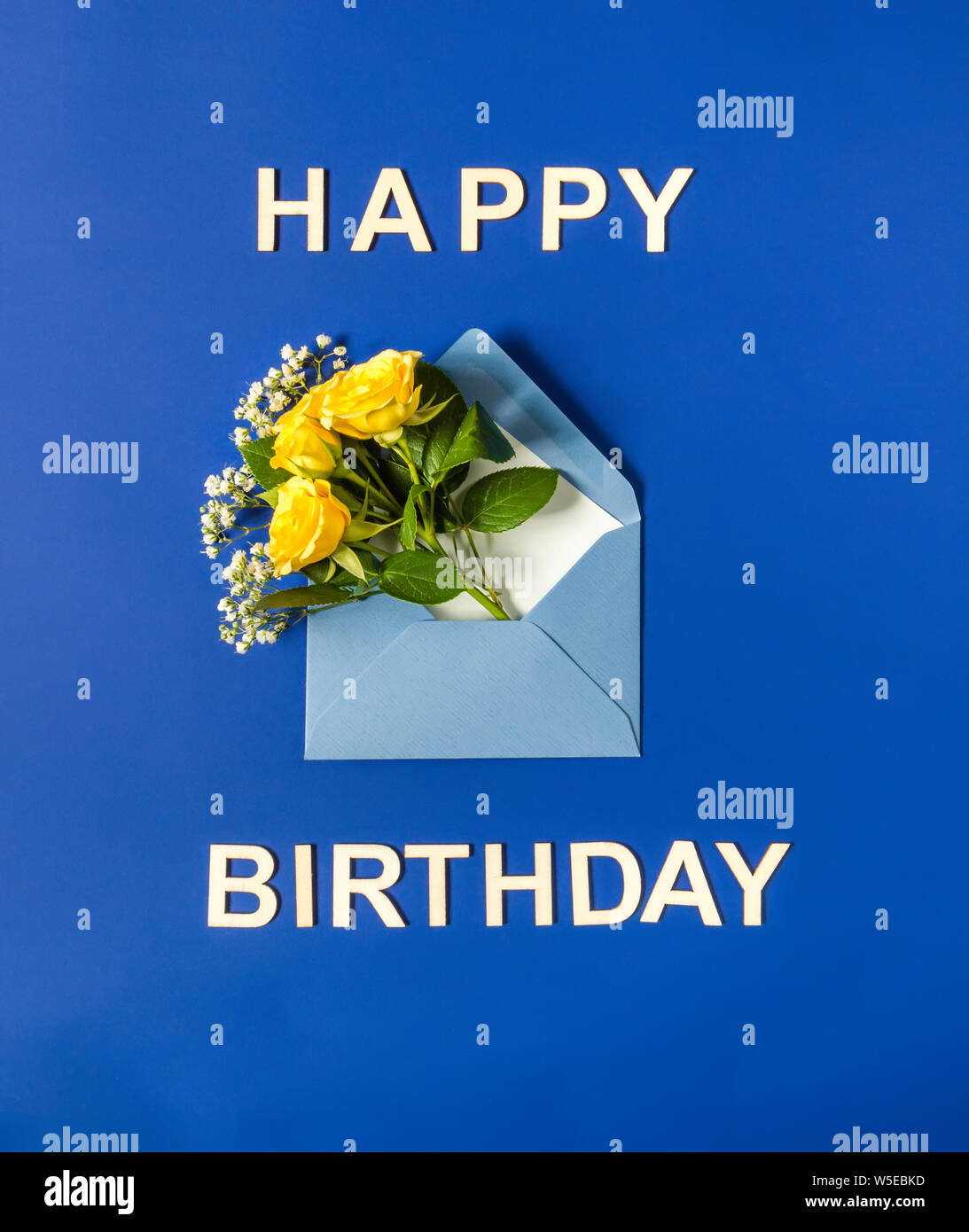 Yellow roses and white Gypsophila in blue envelope close-up on blue background. Text Happy Birthday, wooden letters. Top view, flat lay. Template for Stock Photo