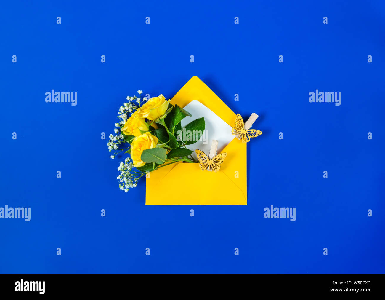 Open envelope with yellow roses, small white filler flowers Gypsophila and yellow butterflies on blue background. Top view, flat lay. Concept of sendi Stock Photo