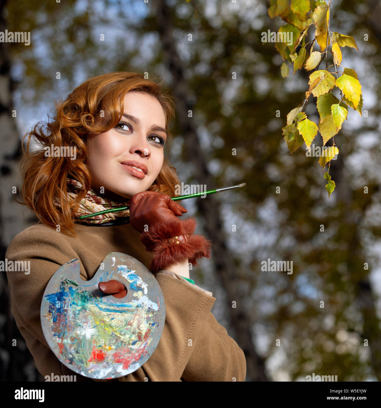 Beautiful woman with palette and brush paints colors leaves on the trees Stock Photo