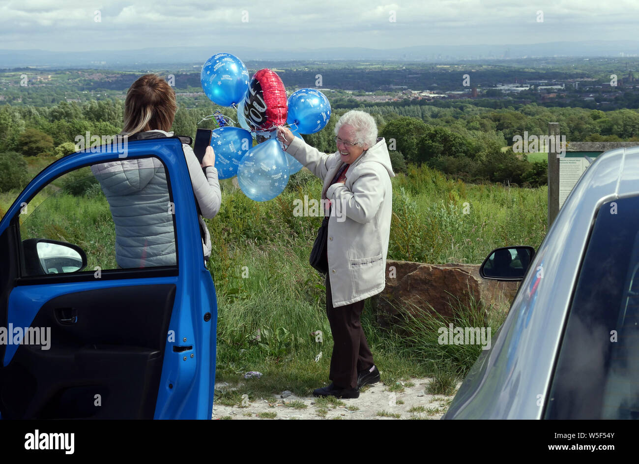 Elderly lady releasing balloons, one balloon is a heart shape saying “Love You” as a young lady records the event with her smart phone on a car park Stock Photo
