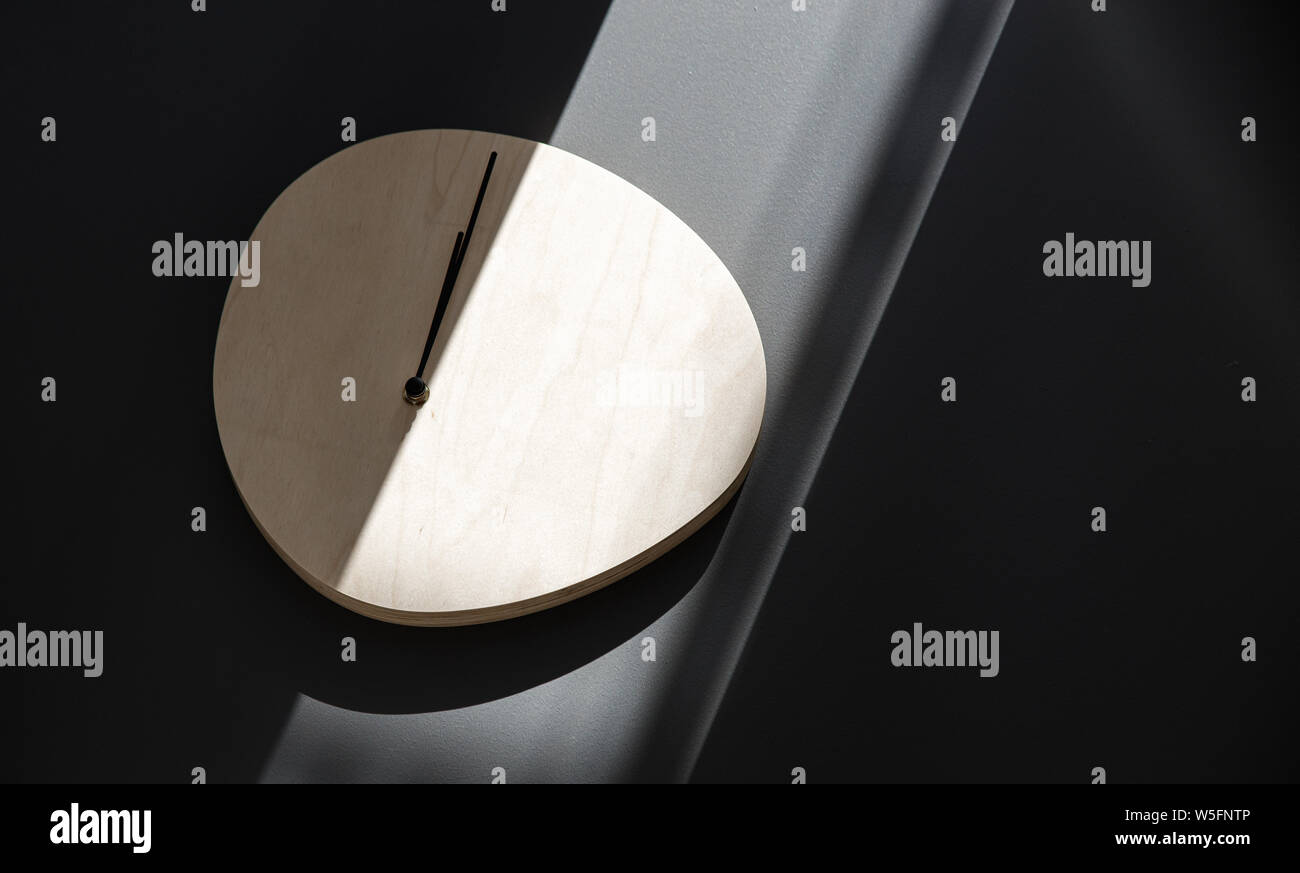 Wooden wall clock hanging on a gray wall in a beam of sunlight. Stock Photo