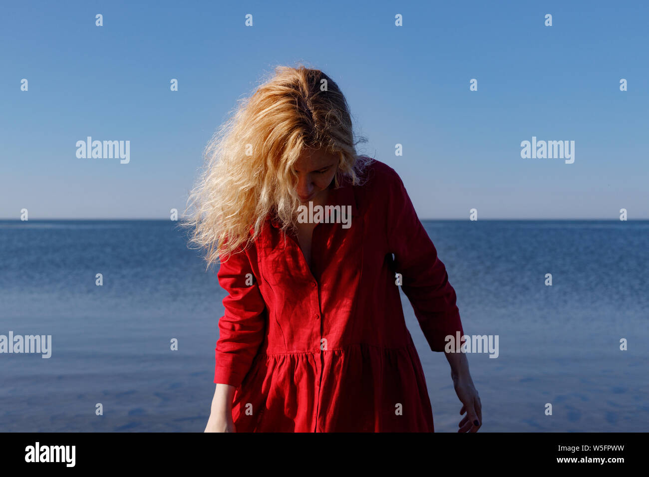 Blonde woman in a red long linen dress standing in the sea in the morning. Stock Photo