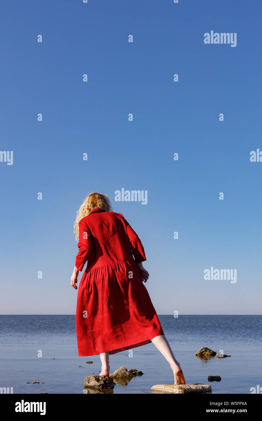 Blonde woman in a red long linen dress standing in the sea in the morning. Stock Photo