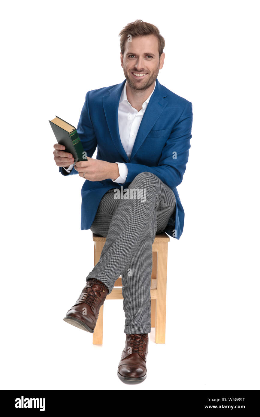 Confident casual guy holding a book and smiling while wearing a suit and sitting on chair on white studio background Stock Photo