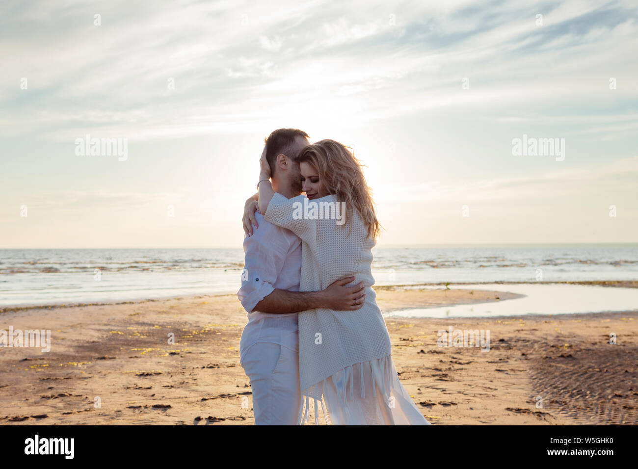 Love, sunset, romance. A young couple in long white soft clothes walking, resting, hugging against the backdrop of a calm sea landscape, sunset. Stock Photo