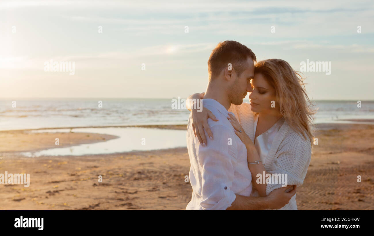 Love, sunset, romance. A young couple in long white soft clothes walking, resting, hugging against the backdrop of a calm sea landscape, sunset. Stock Photo
