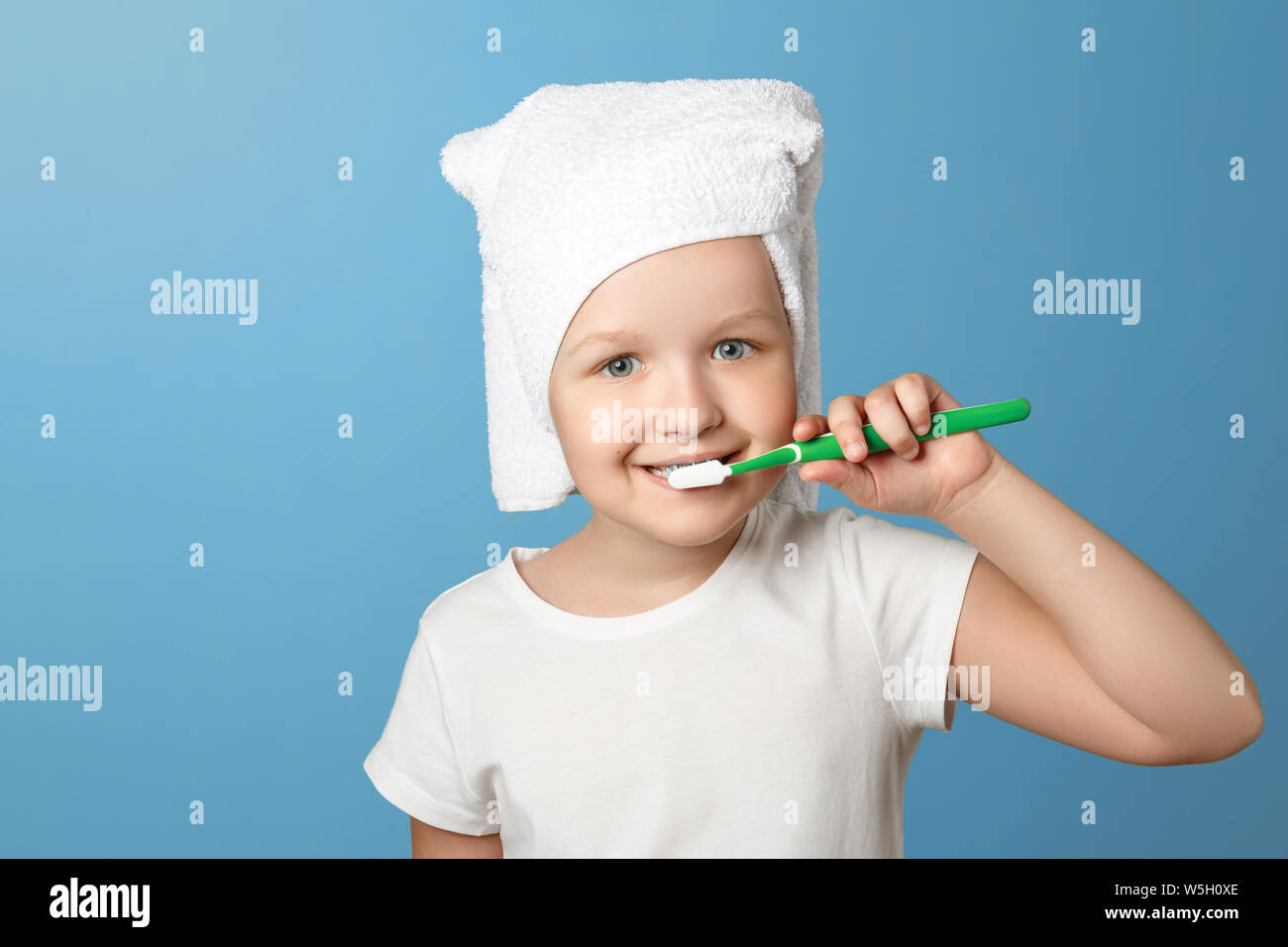Closeup portrait of a little girl on a blue background. A child with a white towel on his head brushing his teeth. The concept of daily hygiene. Stock Photo