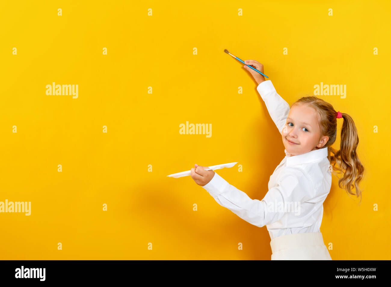 Little girl holding a palette and brush. A child is painting on a yellow empty wall. Stock Photo