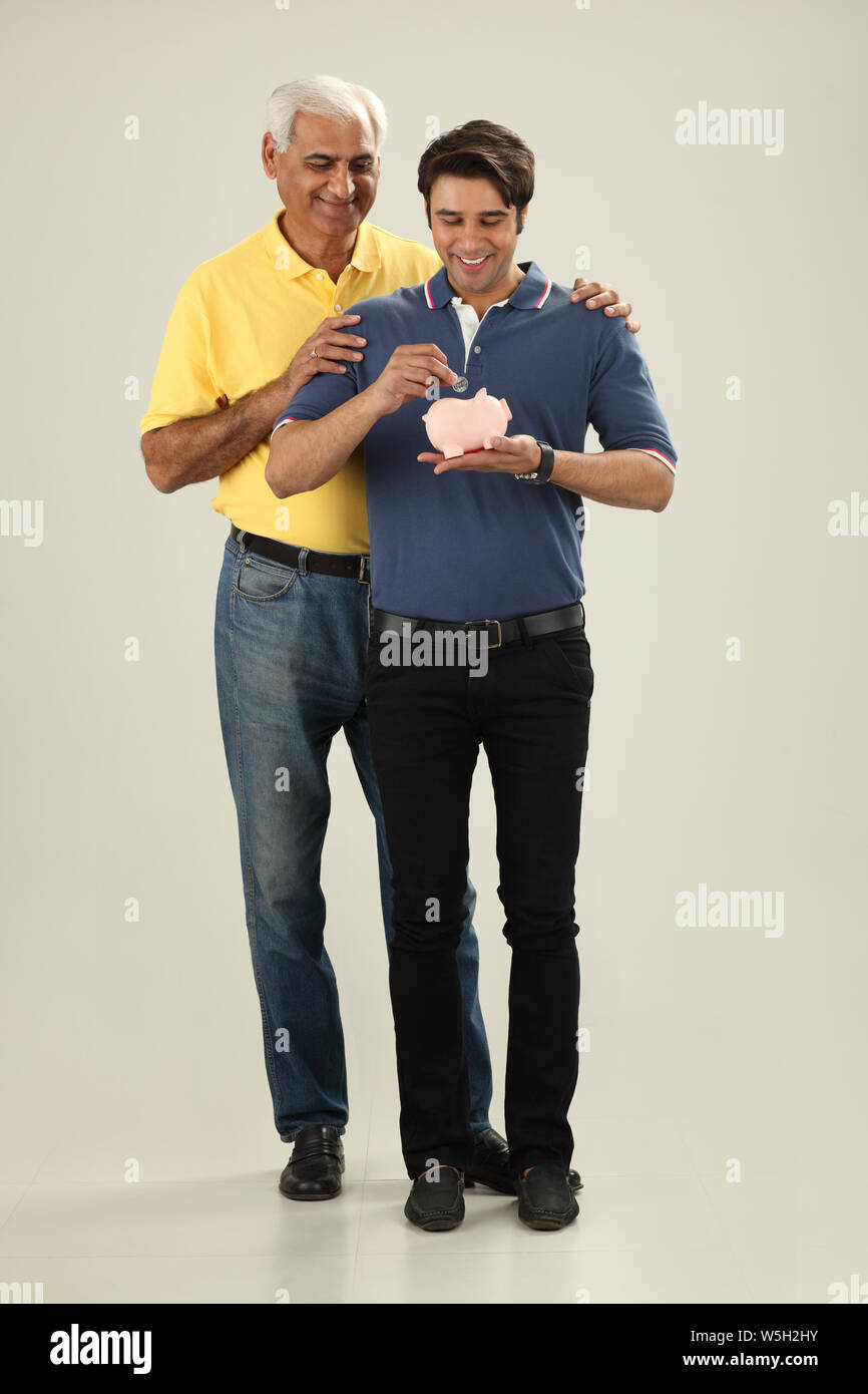 Man putting coin into a piggy bank with his father Stock Photo - Alamy