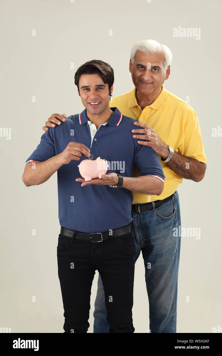 Man putting coin into a piggy bank with his father Stock Photo - Alamy