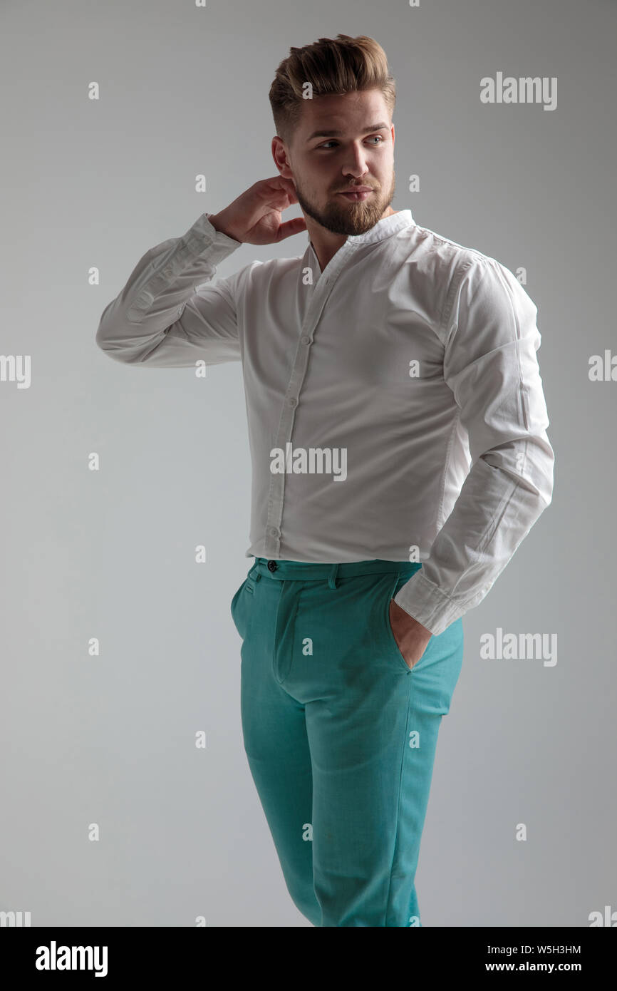 Curious Man Looking Over His Shoulder And Posing With One Hand On His Neck And The Other In His Pocket While Wearing A White Shirt And Standing On Gra Stock Photo