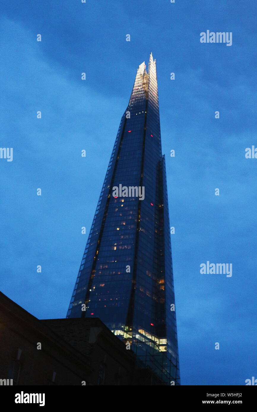 Shard in London, UK in a bluish colour and lights in the windows shot in an low-angle perspective. The photo was taken at night and on a cloudy day. Stock Photo