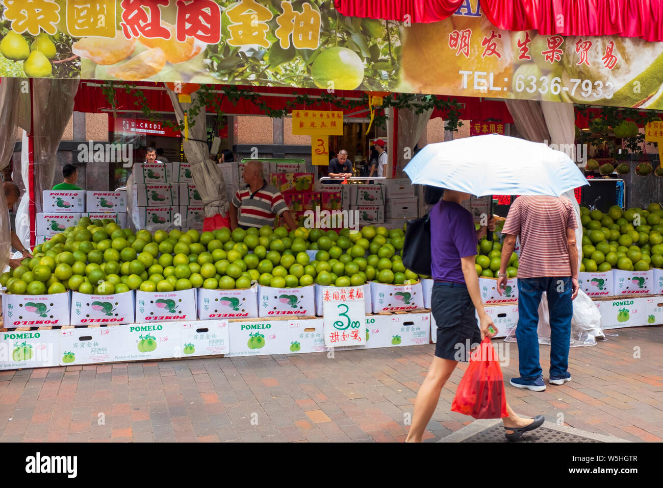 Woman with umbrella walking past stall of green melons for sale at street market in Waterloo St, Singapore Stock Photo