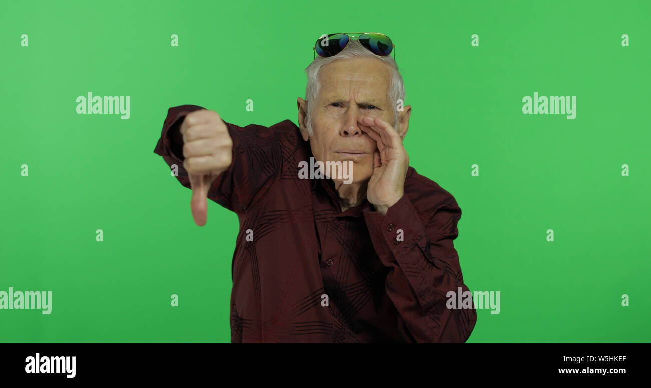 Senior man something giving thumb down. Handsome old man showing displeasure gesture on chroma key background. Elderly grandfather in maroon shirt. Green screen background Stock Photo